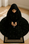 Person wearing a black hijab and dress with white embellishments, sitting on a rug.